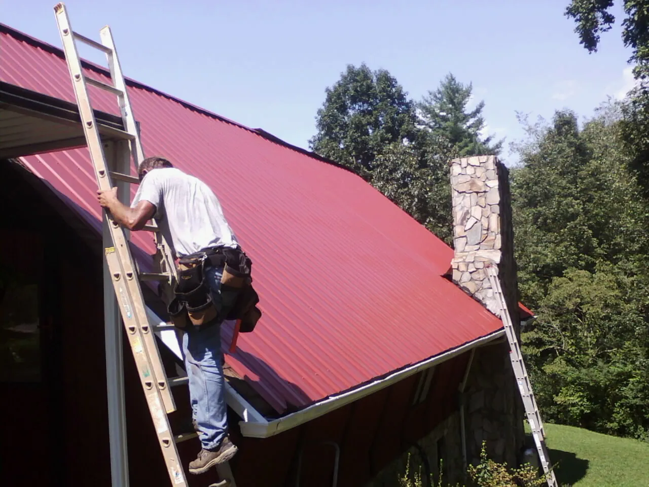 Roofer on a ladder beside a red metal roof and stone chimney — Soffit & Fascia Repair by Roofing Brothers in New Lebanon